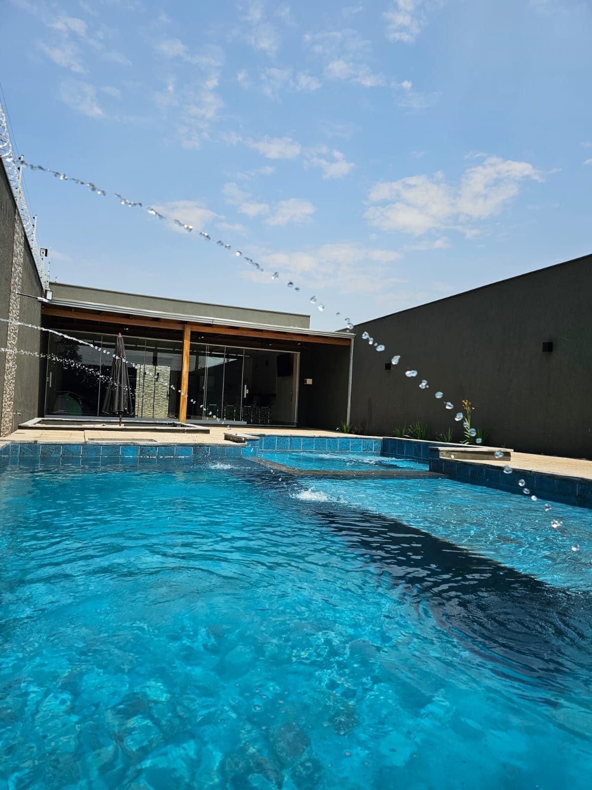 A clear blue pool is visible, surrounded by concrete walls. Water is seen cascading from above, creating a playful atmosphere. Sunlight reflects off the water's surface, and a small area of greenery is observed at the pool's edge, contributing to a calm and refreshing environment.