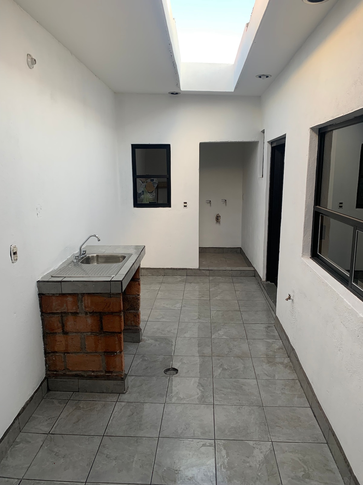 An elongated hallway features a sleek tile floor and white walls, illuminated by natural light from the skylight above. A simple sink is set against a stone base, and two large windows provide views into the exterior. The space is uncluttered and designed for functionality.