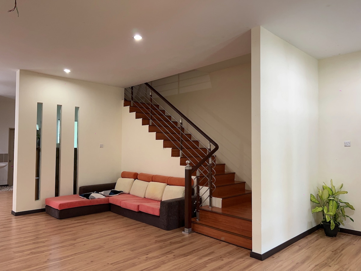 A spacious living area features a sectional sofa in neutral tones, complemented by large mirrors along one wall. A staircase with wooden steps ascends to a second level, while natural light illuminates the room from overhead fixtures.