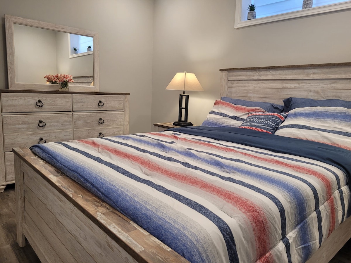 A well-appointed bedroom features a spacious bed with a striped blanket in red, white, and blue hues. A wooden dresser and mirror are positioned nearby, with a lamp providing soft illumination. Natural light filters through a window above the headboard.
