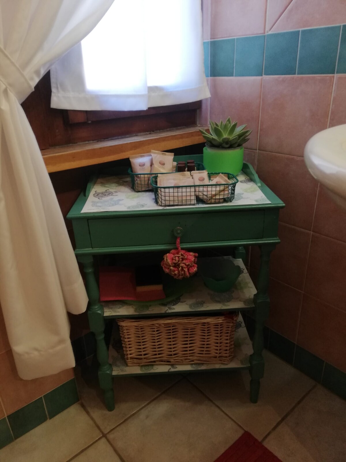 A green nightstand stands in the bathroom, featuring a basket with toiletries and a decorative plant. The piece includes two shelves, one with a woven basket, while natural light filters through a nearby window dressed in white curtains.
