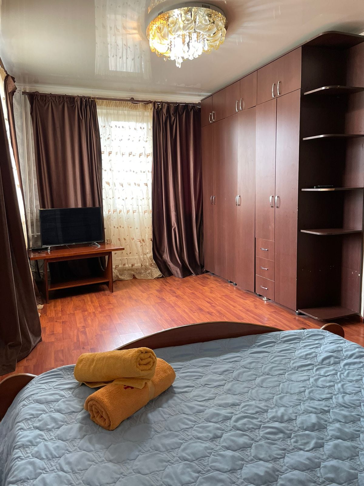 A spacious bedroom features warm wooden flooring. A comfortable bed is layered with a light quilt, while neatly rolled towels are placed on top. A tall wardrobe and shelving unit provide ample storage. Natural light filters through the brown curtains, enhancing the cozy ambiance.
