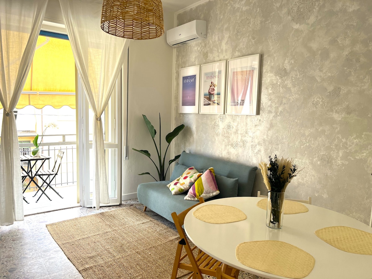 A bright living area features a light blue sofa adorned with colorful pillows. A round white dining table is set with placemats. Large windows allow natural light to fill the space, while a small balcony is visible in the background, framed by flowing curtains and a potted plant.