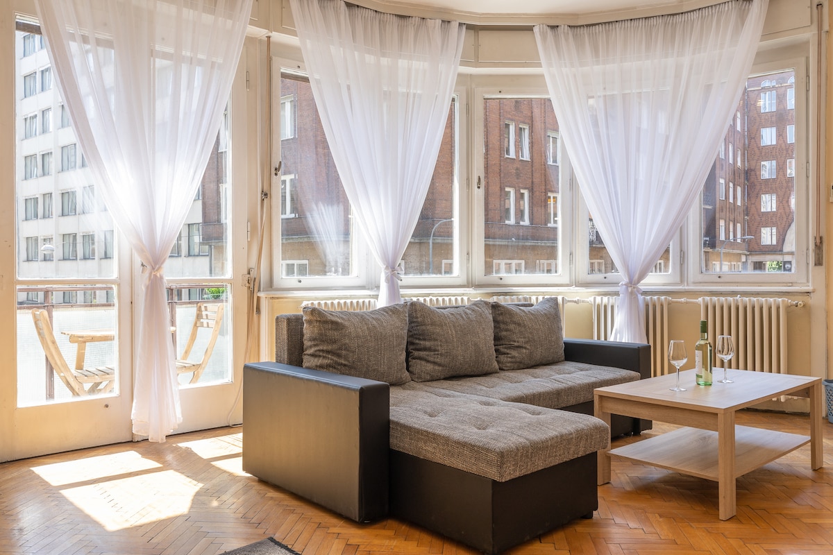 A bright living space is filled with natural light, featuring a modern couch paired with a simple wooden coffee table. Sheer curtains frame the windows, creating a soft ambiance. The room showcases parquet flooring and views of the surrounding urban landscape.