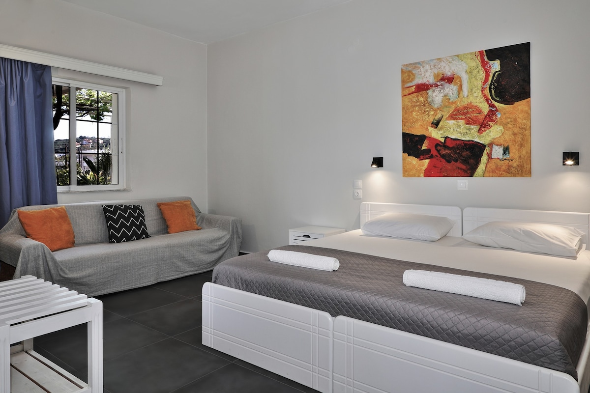 A bright and airy room features a spacious bed with crisp white linens. A gray sofa adds comfort, with orange and patterned cushions providing color. A decorative painting is displayed above the bed, and natural light filters in through a window with blue curtains.