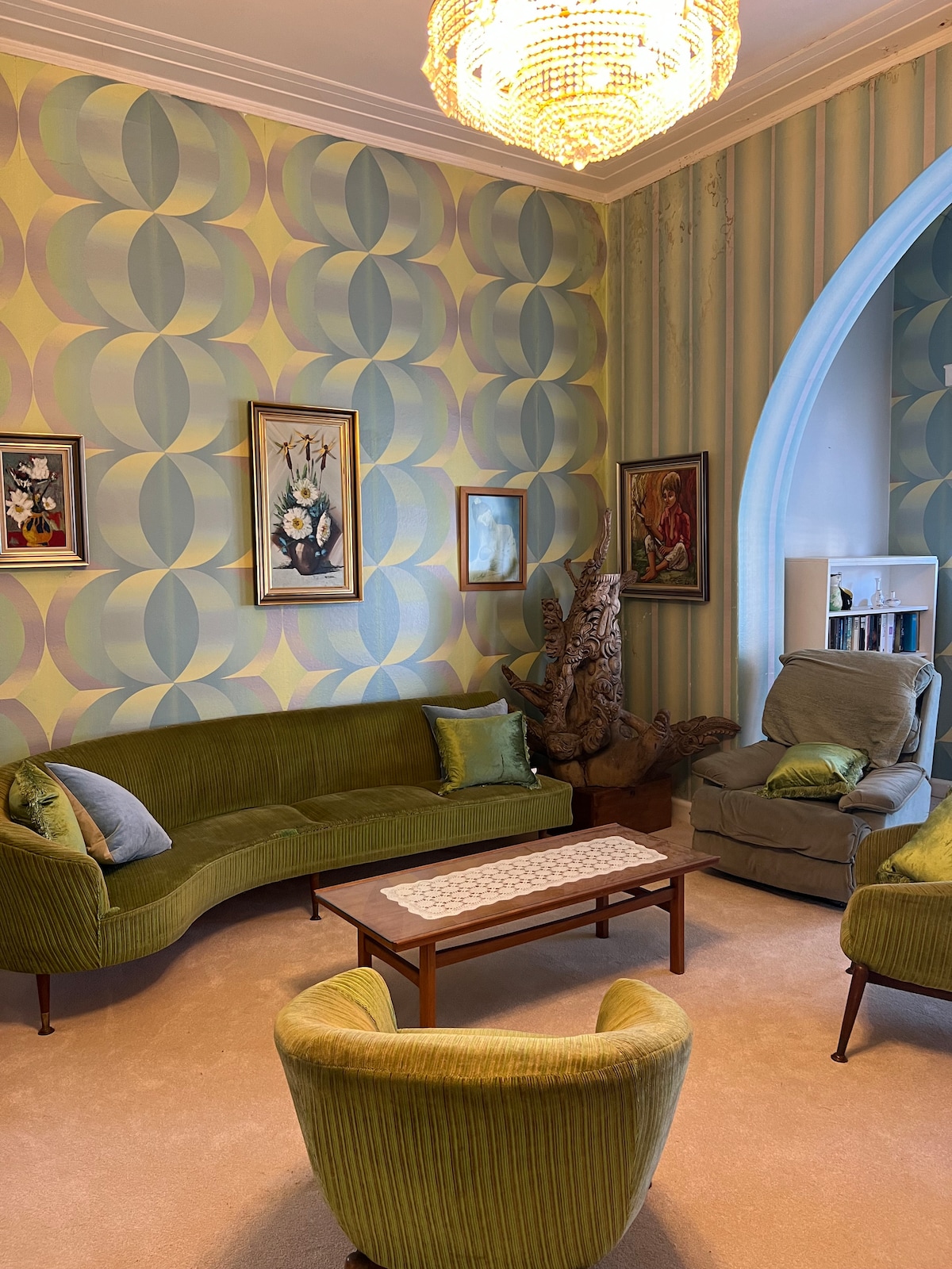 A spacious lounge area features retro furnishings in vibrant green. Textured wallpaper with a geometric pattern adorns the walls, while framed artwork adds a personal touch. A decorative sculpture complements the setting, and a wooden coffee table sits centrally, inviting relaxation.