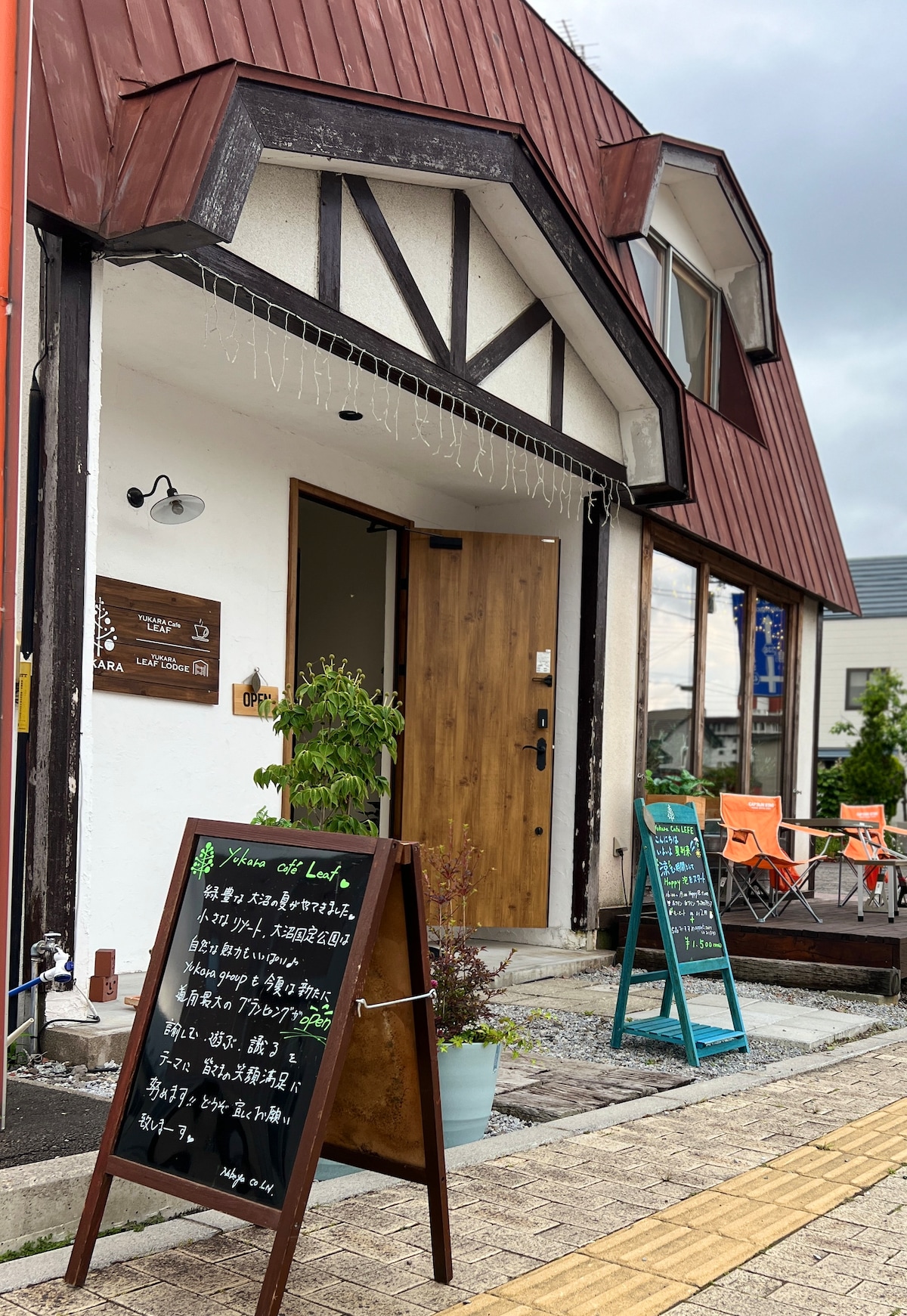 The exterior of a guesthouse features a wooden door framed by a distinct roofline. Nearby, two chalkboards display information. Potted plants add greenery, and colorful chairs are visible on a patio, inviting guests to relax outdoors.