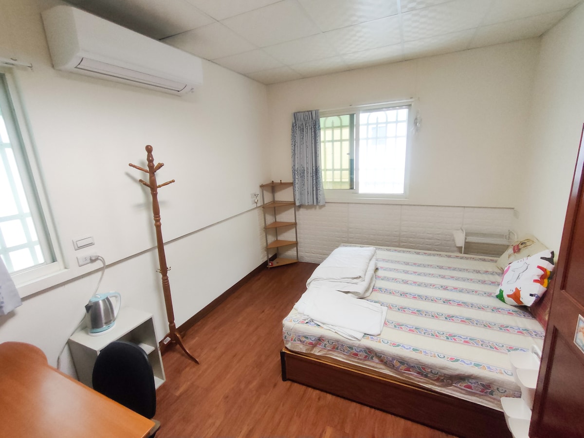 A bright room features a spacious bed with fresh linens and a lightweight blanket. A wooden desk and chair are positioned near a window, allowing natural light to fill the space. A clothing rack stands beside a shelf, providing convenience and organization.