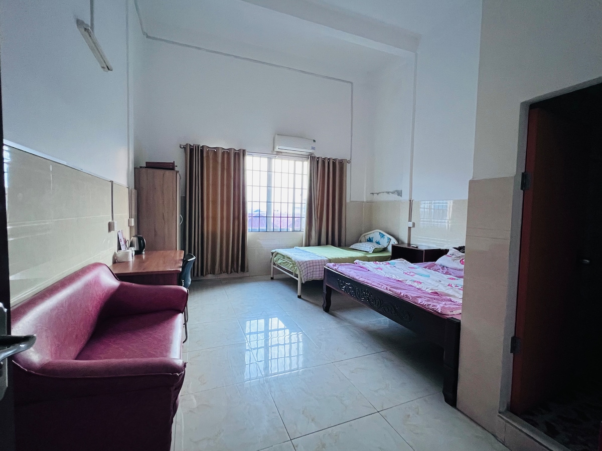 A spacious room features two beds, one adorned with a floral blanket and the other with a simple cover. A light-colored sofa is positioned against one wall, while a desk and chair are visible nearby. Natural light streams in through the large window, illuminating the space.