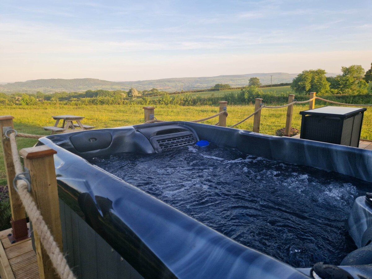 An outdoor hot tub is positioned on a wooden deck, offering a view of rolling hills and fields in the distance. The water is calm, reflecting the clear sky above, while wooden seating can be seen nearby, surrounded by natural greenery.