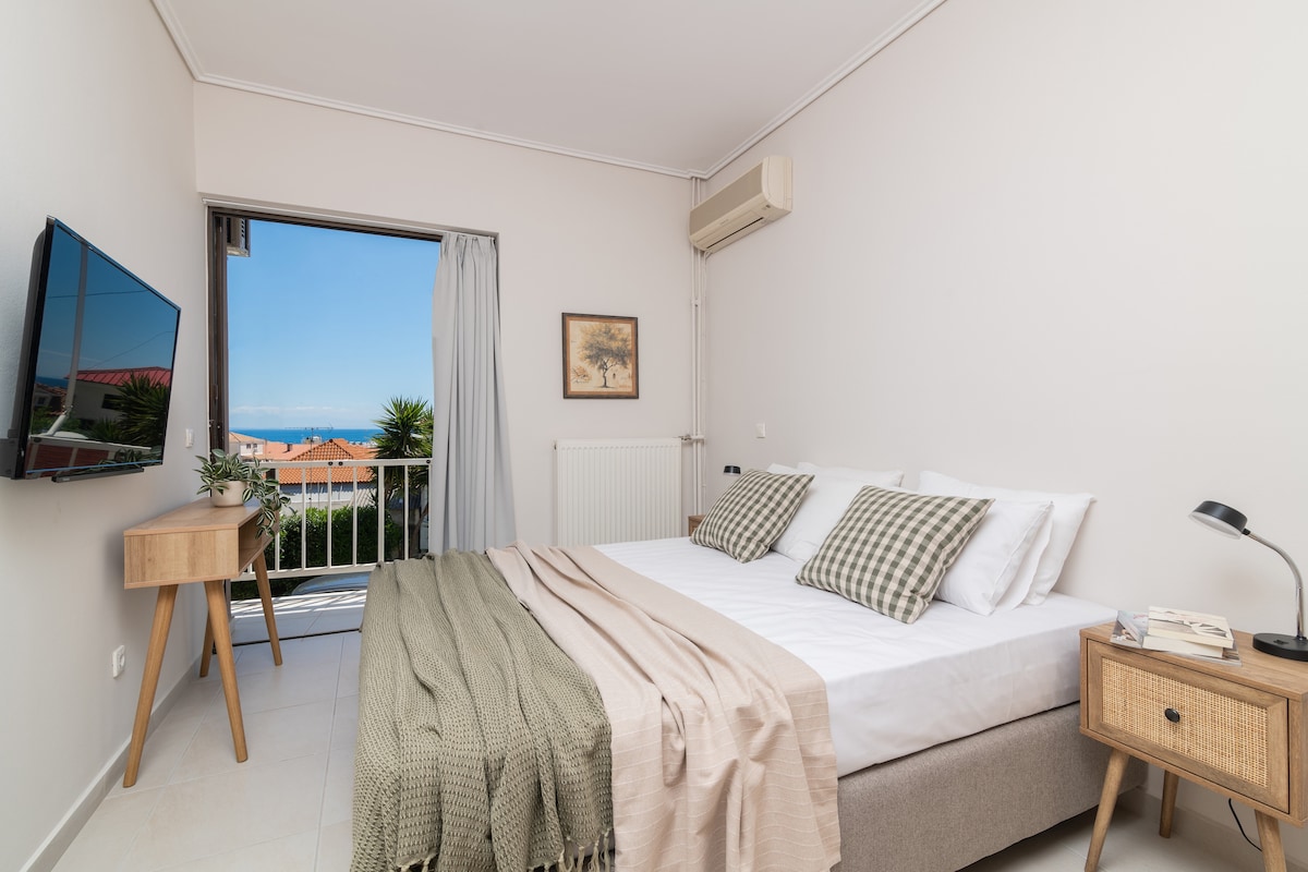 A bright bedroom is arranged with a spacious bed dressed in neutral linens and plaid throw pillows. A small side table holds a book and lamp. A large window opens to a view of greenery and distant sea, with natural light illuminating the room.