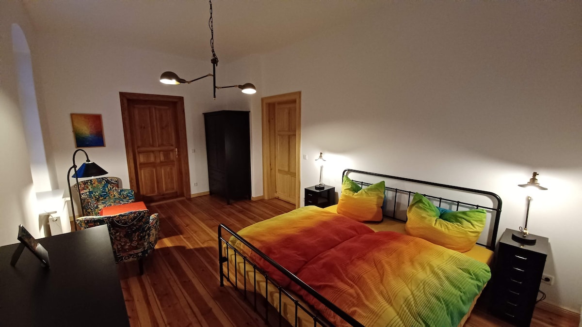 A vibrant bedroom features a large bed with colorful bedding in shades of green and yellow. A cozy armchair is situated next to a desk, while warm wooden flooring complements the neutral-colored walls. Two bedside tables with lamps add functionality to the space.