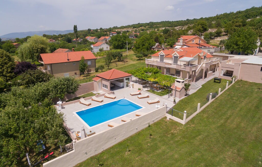 A large outdoor swimming pool is surrounded by lounge chairs and shaded areas, set within a spacious garden. The property features well-maintained landscaping, with residential buildings and greenery visible in the background, contributing to a serene environment.