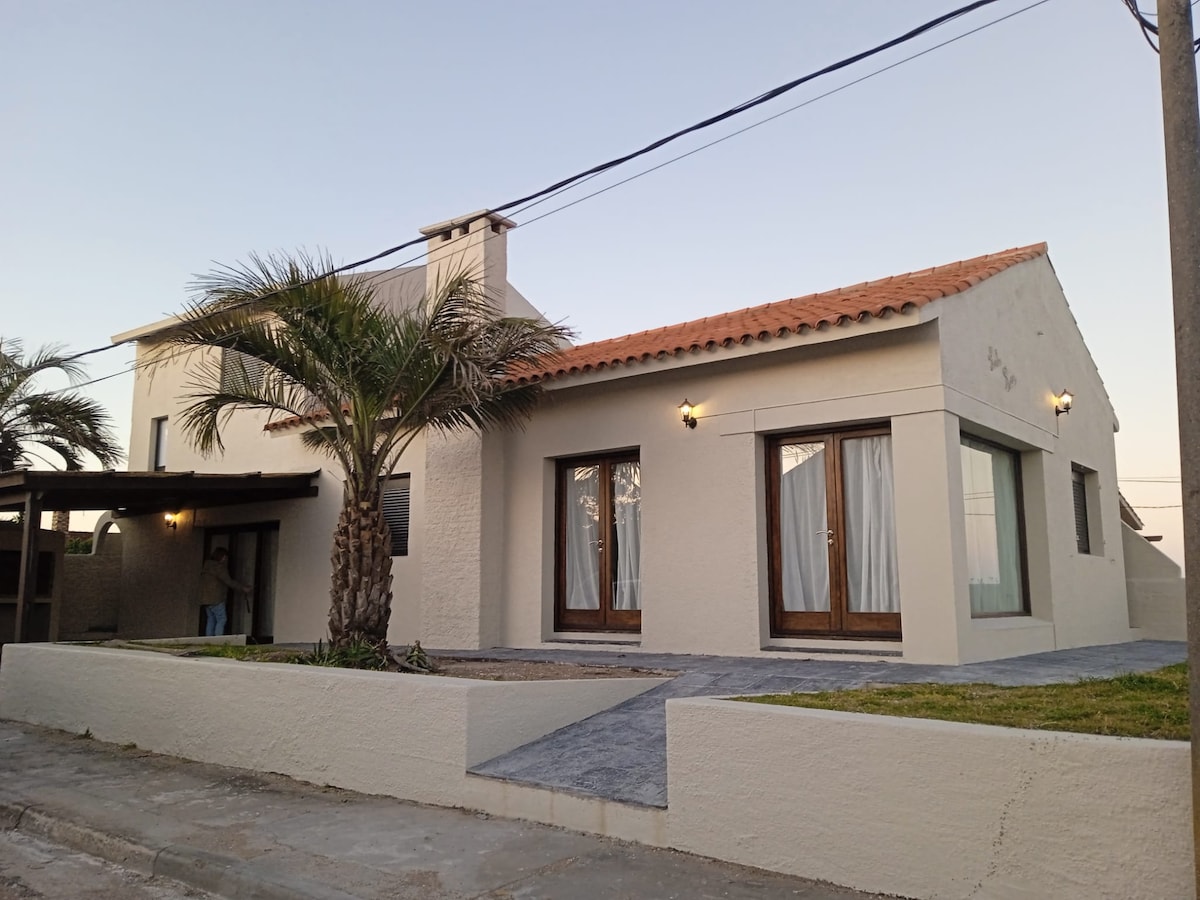 A two-story house is presented with a cream-colored exterior and a red-tiled roof. Flanking palm trees provide a tropical feel, while large windows allow natural light to brighten the interior. An inviting stone pathway leads to the entrance, enhancing the home's curb appeal.