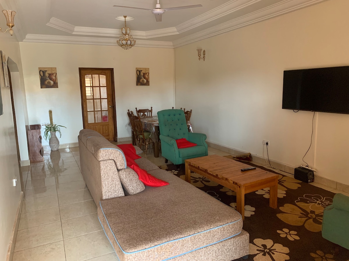 A spacious living area features a large sectional sofa with vibrant red cushions and an accent chair. A wooden coffee table occupies the center, while a wall-mounted television stands across from the seating. A dining table is visible in the background, set to accommodate multiple guests.