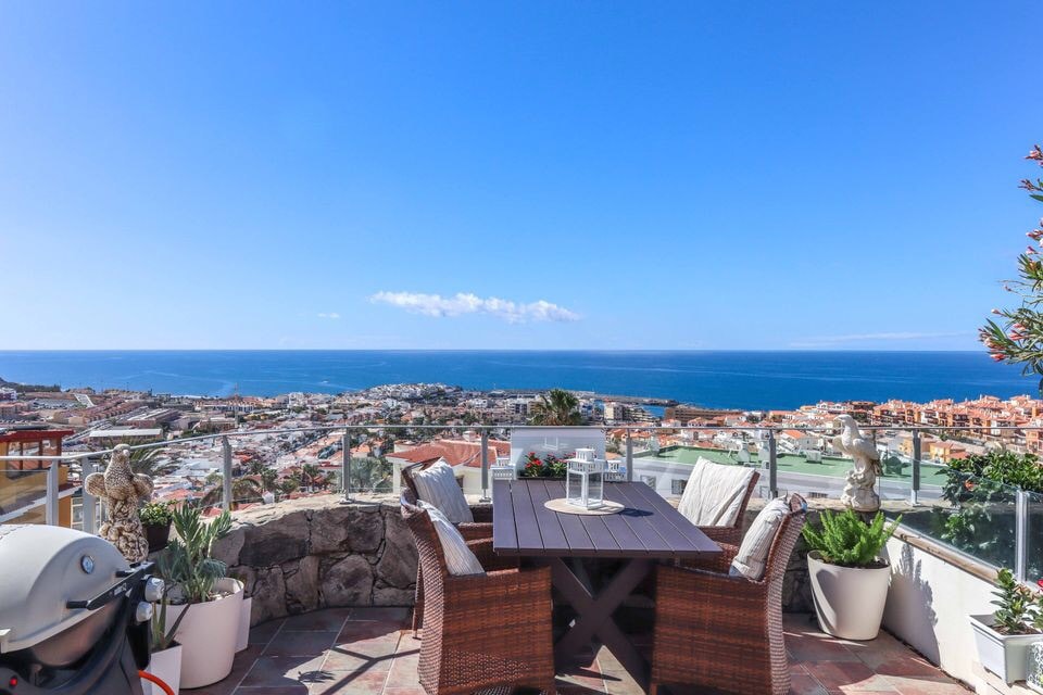 A dining area is set on a balcony, featuring a dark wooden table surrounded by woven chairs. Lush potted plants are positioned around the space. A clear view of the ocean and distant coastal landscape is visible beyond a glass railing, with bright blue skies above.