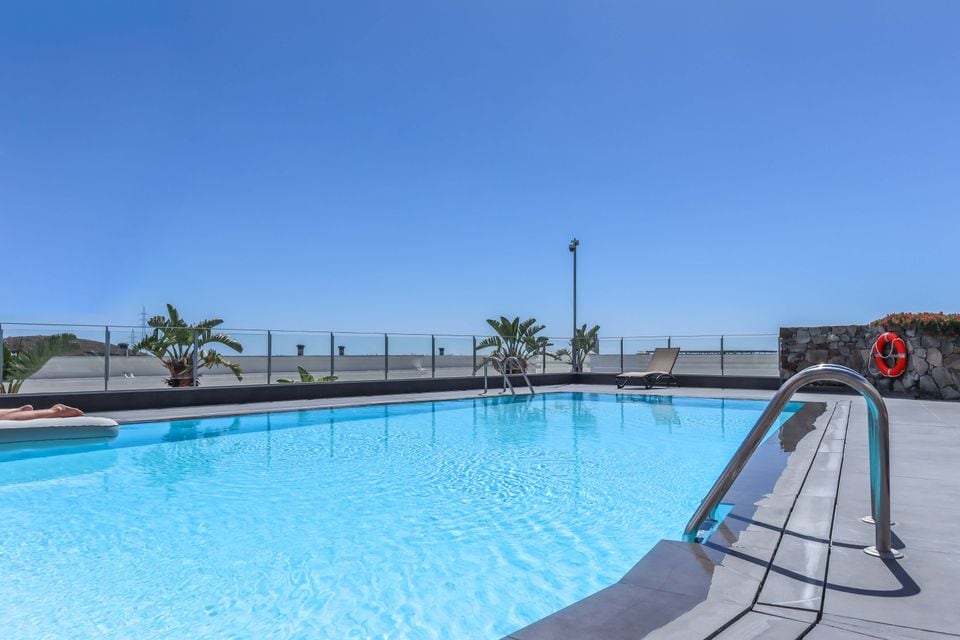 A refreshing outdoor pool is presented with clear blue water, surrounded by a deck featuring lounge chairs. Lush plants are visible around the pool area. The unobstructed view of the horizon is enhanced by a cloudless sky, creating a peaceful and open ambiance.