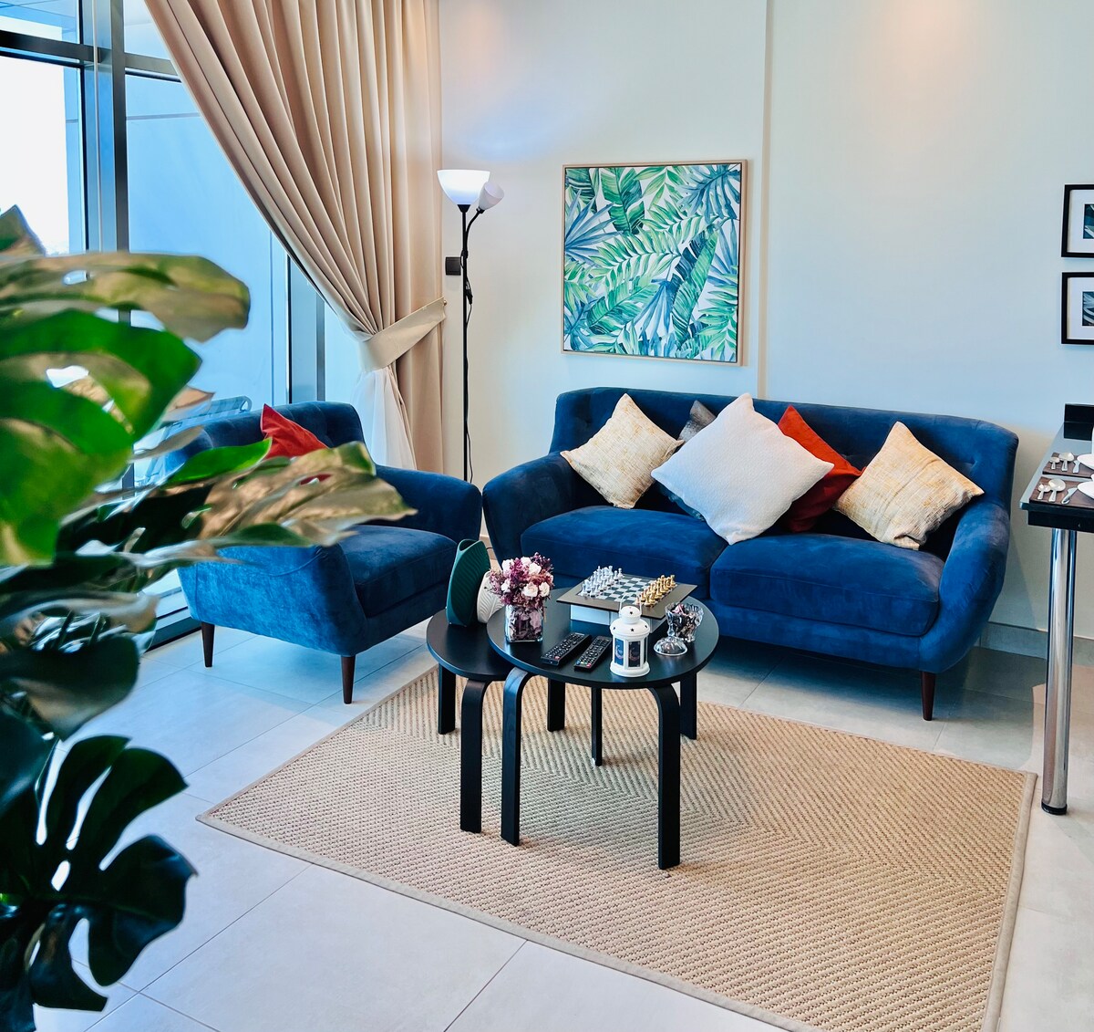 A modern living area is presented with blue upholstered sofas, adorned with decorative cushions in various shades. A round black coffee table sits at the center, accompanied by a small floral arrangement. Natural light streams in through large glass doors, enhancing the open, airy feel.