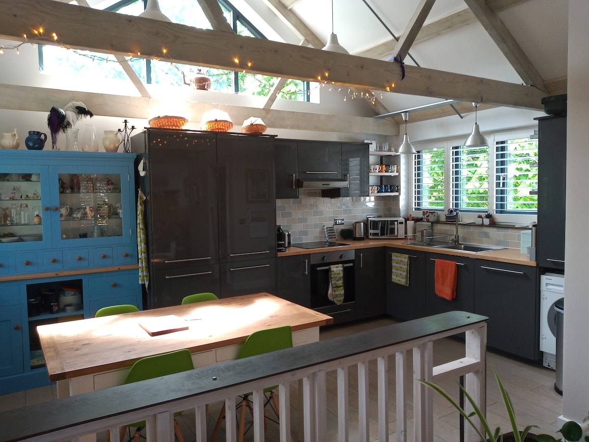 The kitchen features modern cabinetry in dark tones, complemented by a light wooden dining table surrounded by bright green chairs. Natural light floods the space through windows adorned with decorative shutters, highlighting various kitchen appliances and decorative items on the shelves.
