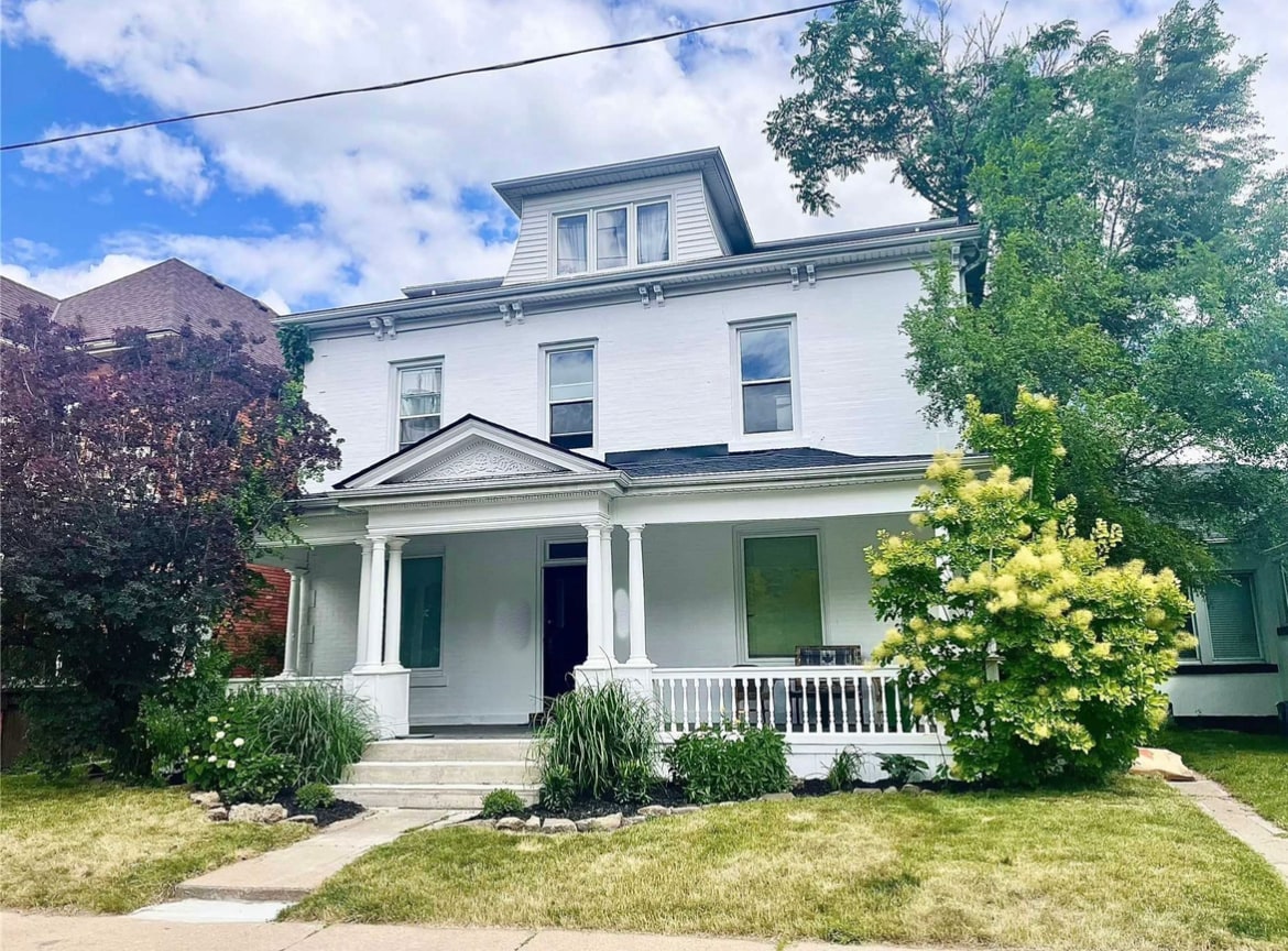The exterior of a fourplex home is displayed, featuring a white façade and a welcoming covered porch. Lush greenery surrounds the property, with trees and shrubs adding to the curb appeal. A well-maintained lawn complements the inviting entrance.