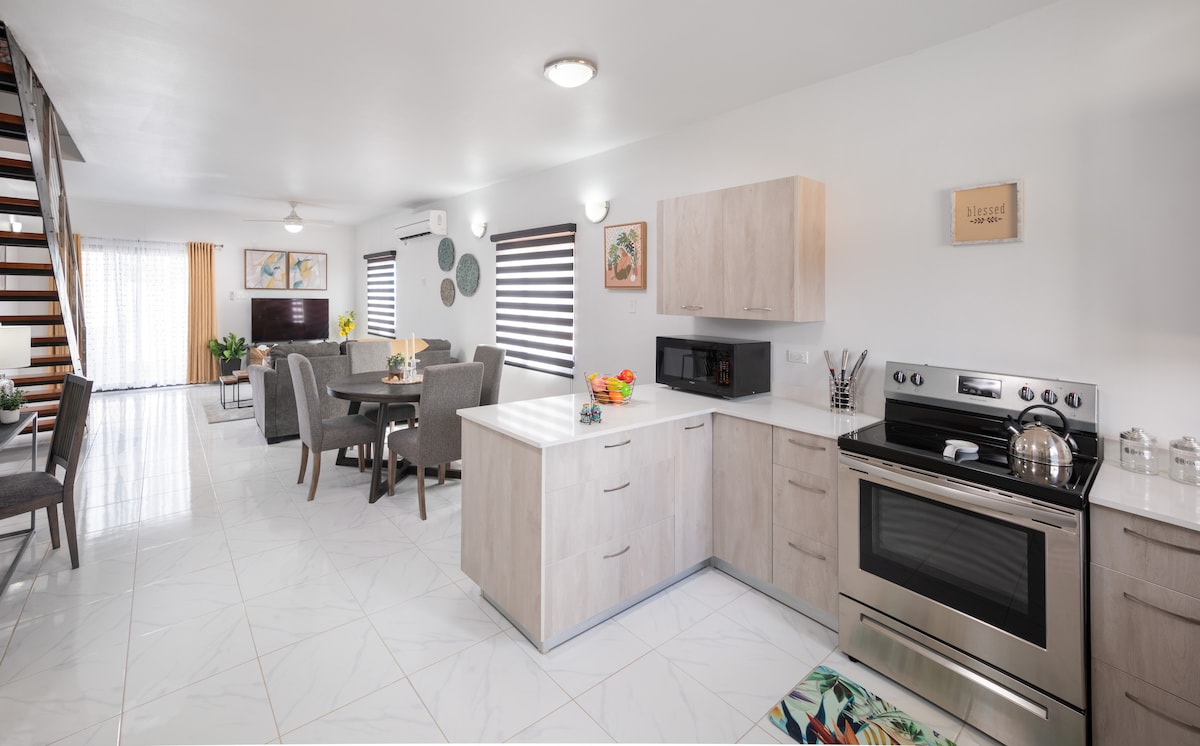 A modern kitchen area features stainless steel appliances, including an oven and microwave, alongside light wood cabinetry. A dining space with gray chairs sits nearby, and large windows allow natural light to fill the open floor plan, enhanced by light-colored flooring.
