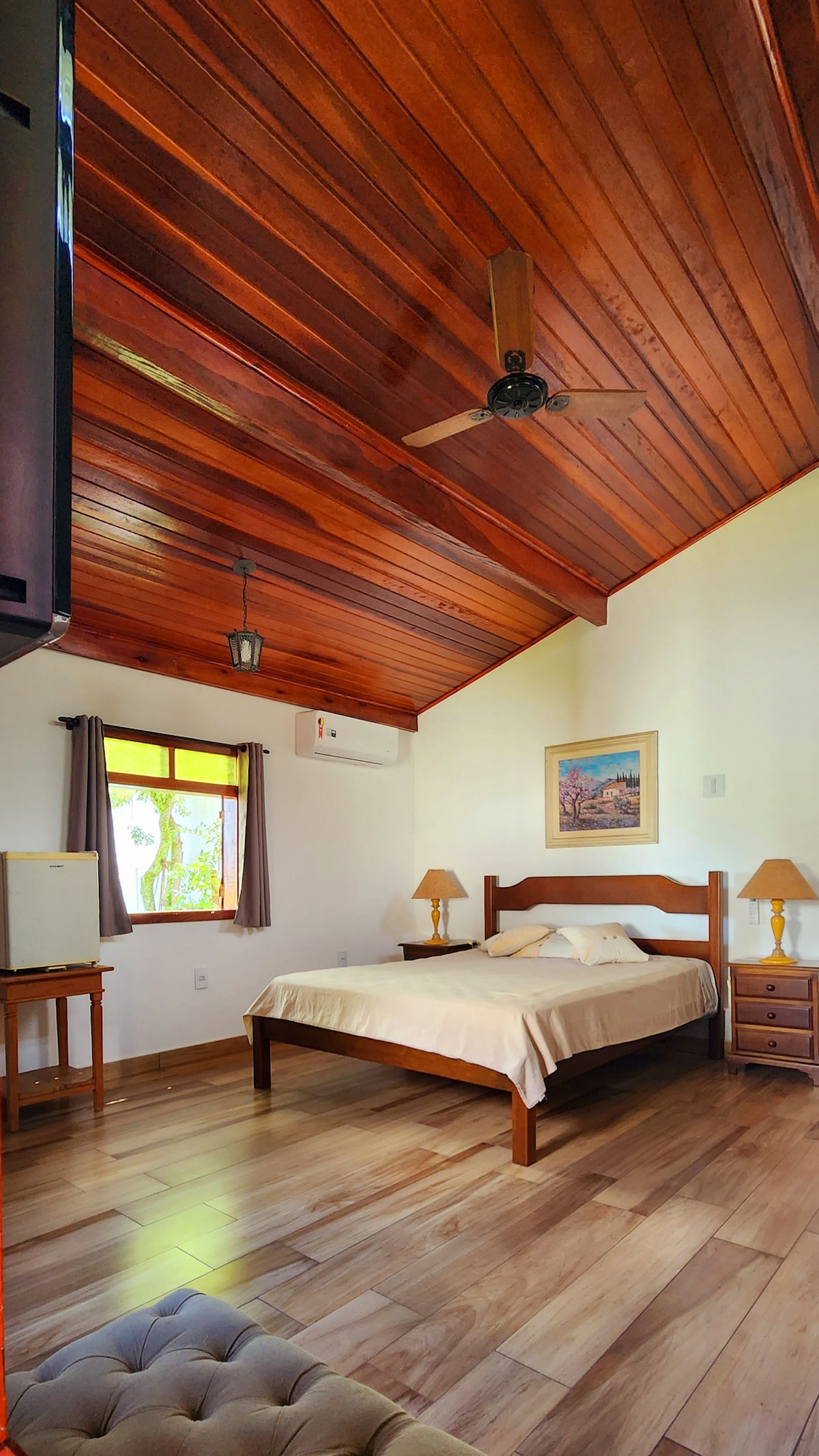 A cozy bedroom features wooden beams on the ceiling and warm-toned walls. Natural light enters through a window with drapes, illuminating a spacious bed, two bedside tables with lamps, and a small writing desk. Decorative artwork adds a personal touch to the inviting space.