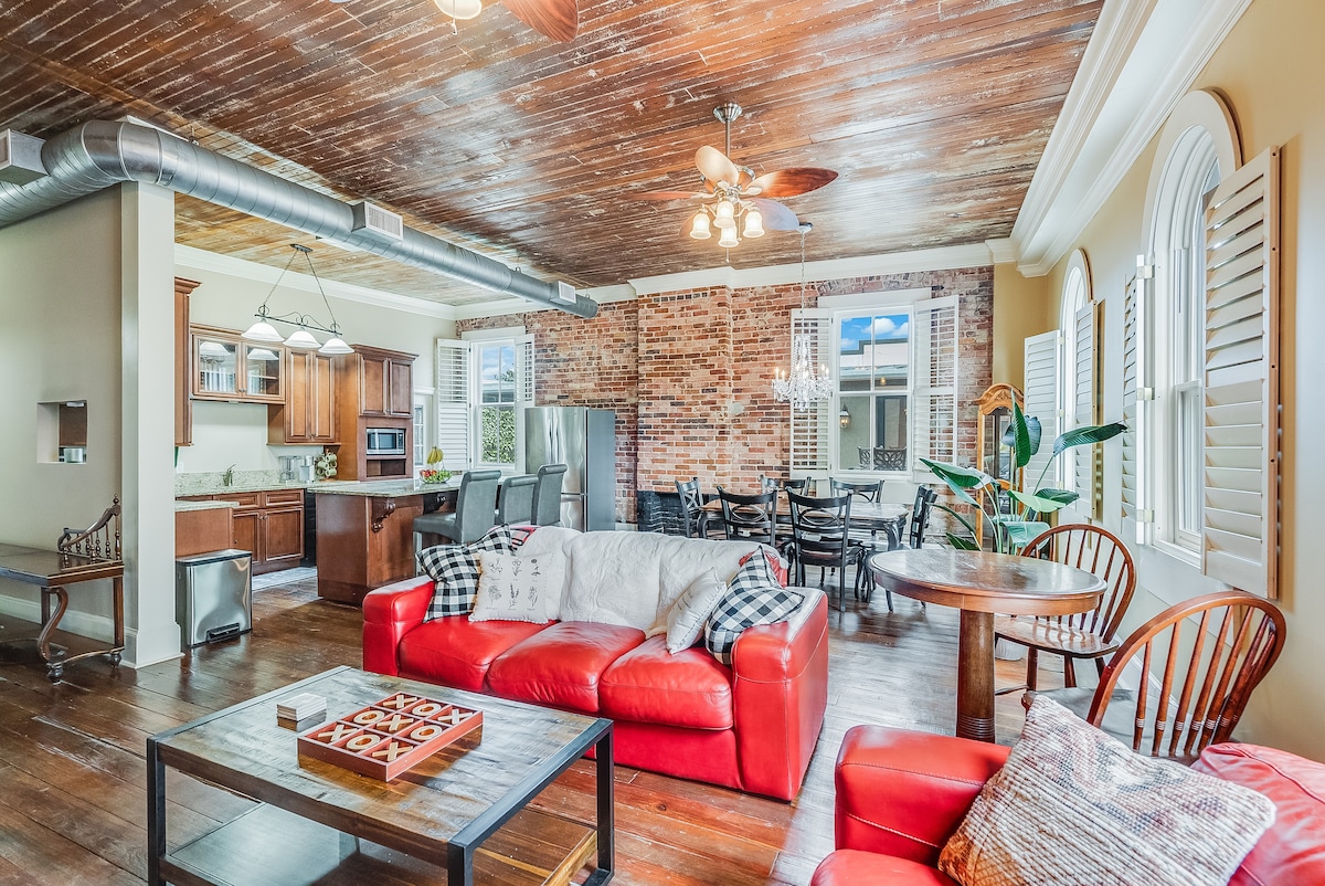 An open-concept living area features hardwood floors and exposed brick walls. A red sectional sofa is positioned with a relaxed throw, while a round dining table is visible near the kitchen. Natural light floods in through large windows and ceiling fans enhance air circulation.