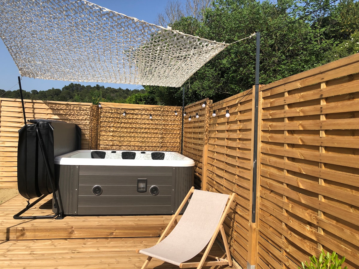 A private outdoor jacuzzi is positioned under a shaded area, surrounded by wooden fencing for privacy. A single lounge chair rests nearby, allowing for relaxation. Soft lighting is incorporated into the setting, complementing the natural greenery in the background.