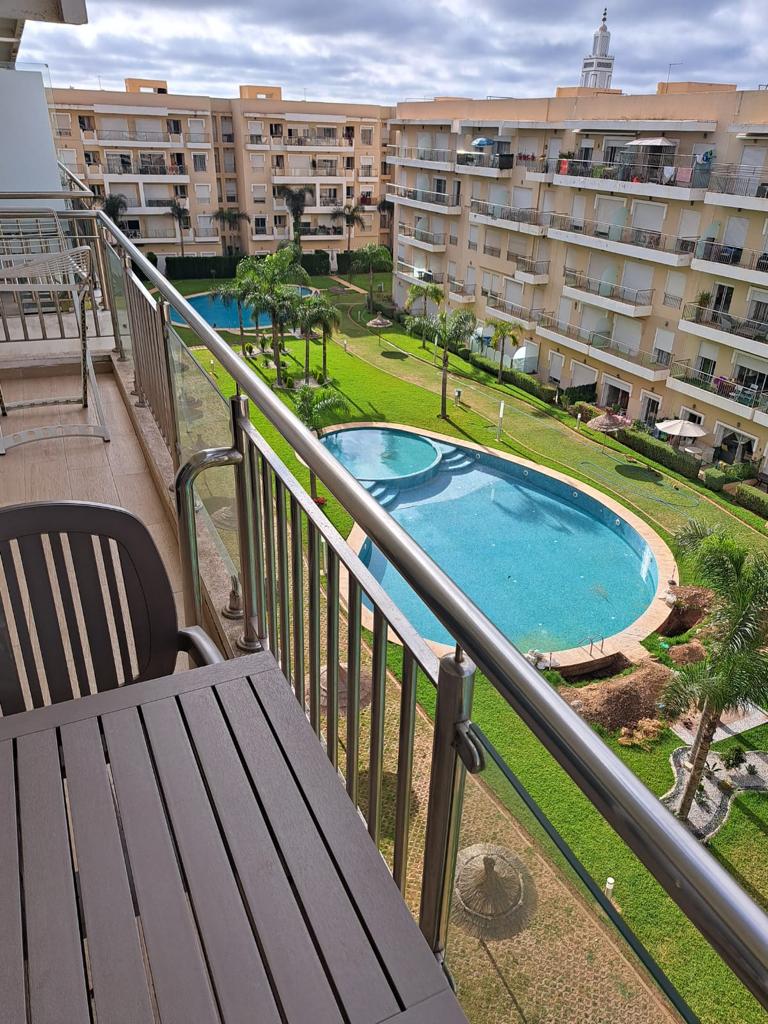A balcony view showcases a serene pool surrounded by well-maintained green areas. Spacious lawns and palm trees are visible, contributing to a tranquil outdoor atmosphere. Nearby residential buildings are seen in the background, enhancing the urban setting.