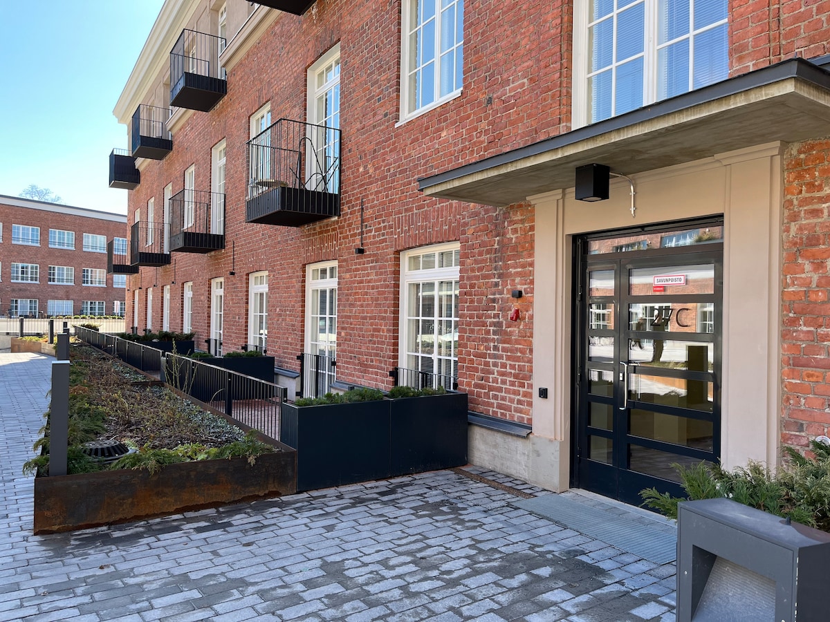 The entrance of a historic building is framed by weathered brick walls and large windows. A set of glass doors is visible, along with several small balconies. The pathway is made of cobblestones, bordered by landscaped greenery and modern planter boxes.