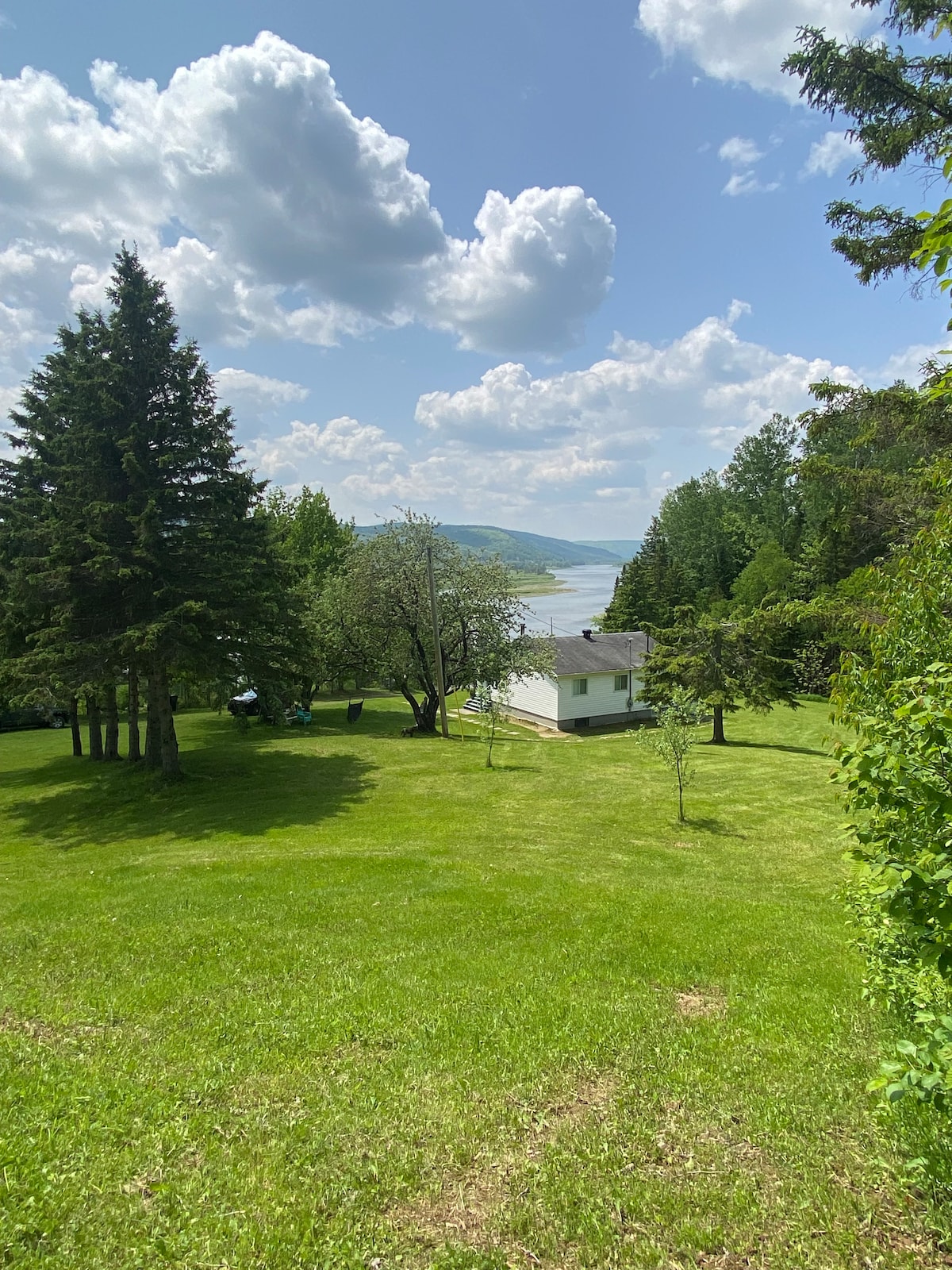 A white cottage is situated on a lush green lawn, surrounded by trees, with a view of the river in the background. The sky is partially cloudy, allowing daylight to illuminate the serene outdoor space.
