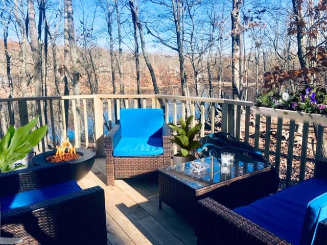 An outdoor deck area features comfortable wicker seating with bright blue cushions, complemented by a coffee table adorned with a few glasses. A fire pit adds warmth, and greenery surrounds the space, providing a serene view of the wooded area beyond.