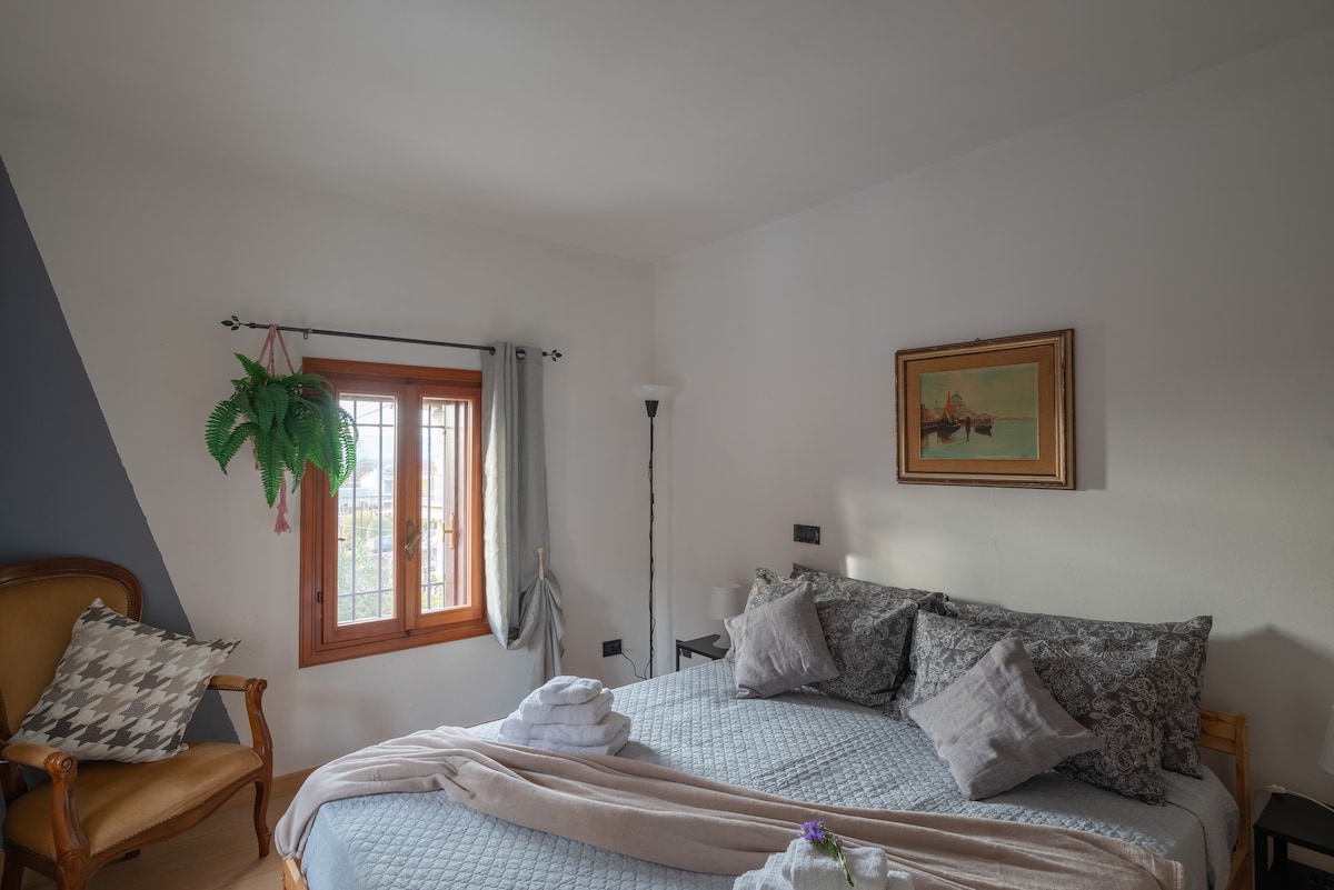 A cozy bedroom features a spacious bed adorned with patterned pillows and a soft blanket. A comfortable chair is set beside a window, which is framed by light curtains. A piece of artwork hangs on the wall, adding a touch of elegance to the room.