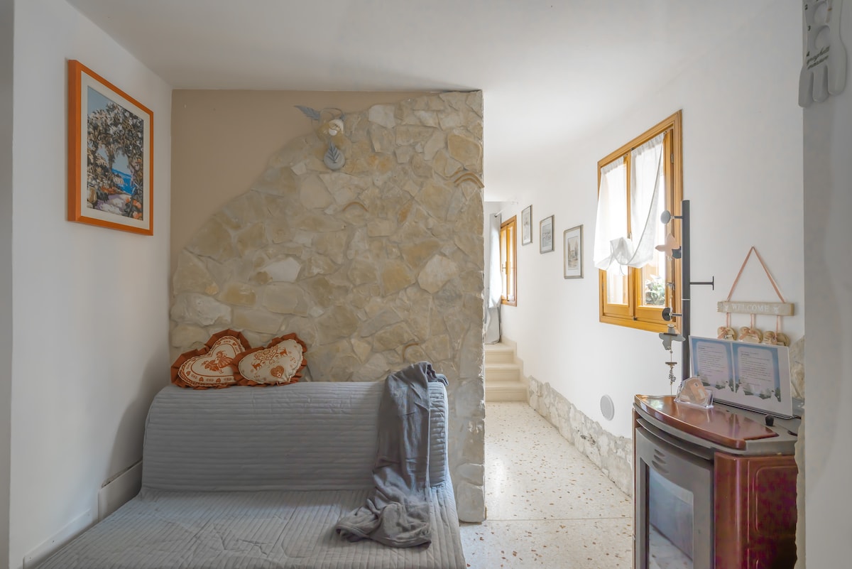 A light-filled hallway features a textured stone wall and a gray sofa adorned with decorative throw pillows. Windows with sheer curtains allow natural light to illuminate the space, while framed pictures add character to the walls. A staircase leads to the upper level.