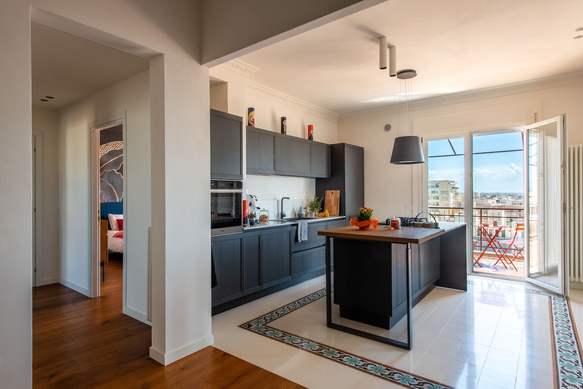 The modern kitchen showcases dark cabinetry and an island with a gas cooktop. Natural light flows in through an open door leading to a balcony. A tiled backsplash adds visual interest, with kitchen essentials neatly arranged on the counter.