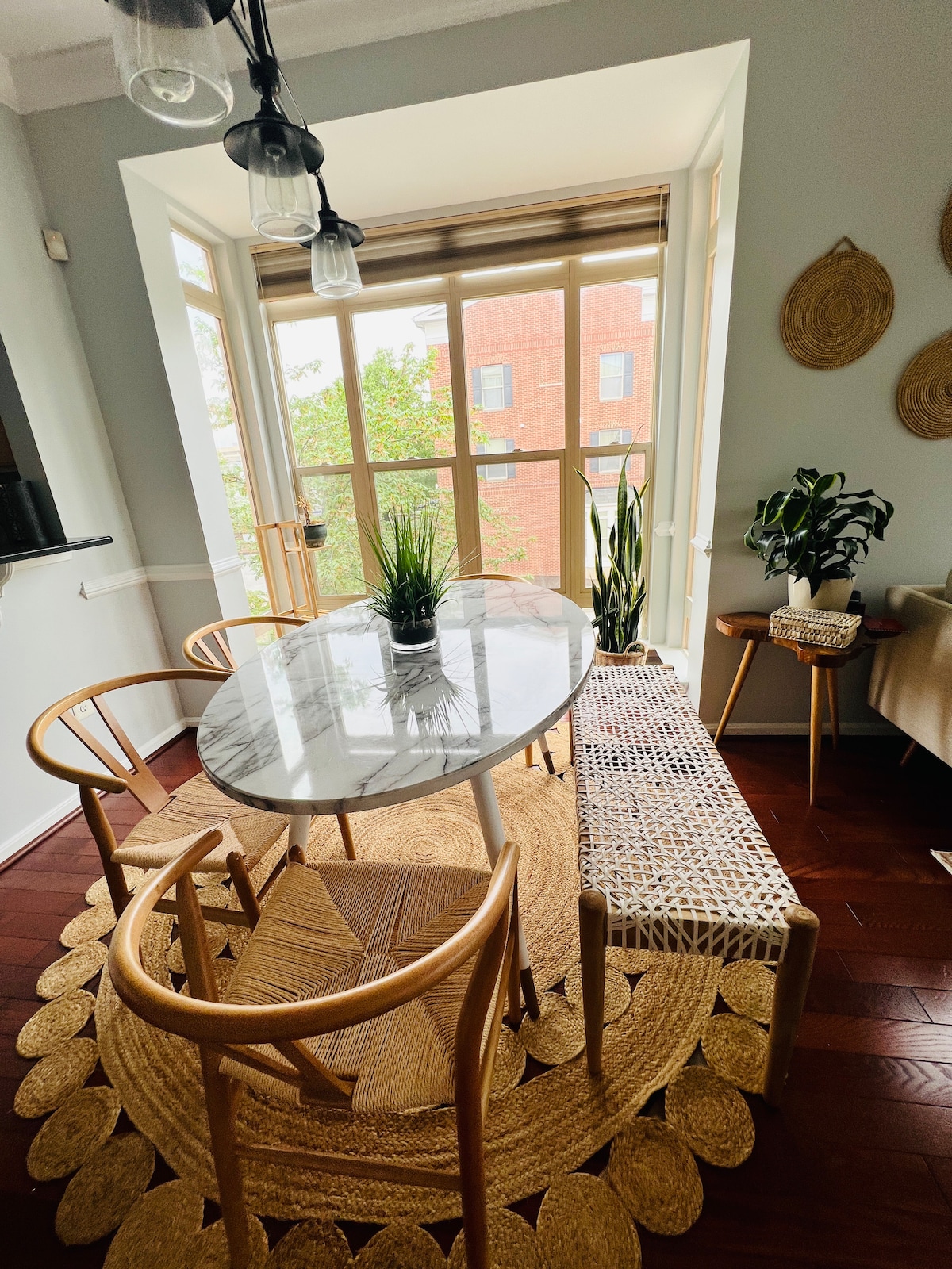 A dining area features a marble-top table surrounded by four wooden chairs and a woven bench. Natural light enters through large windows adorned with beige treatments. Green plants add a touch of life, complemented by a circular jute rug that anchors the space.