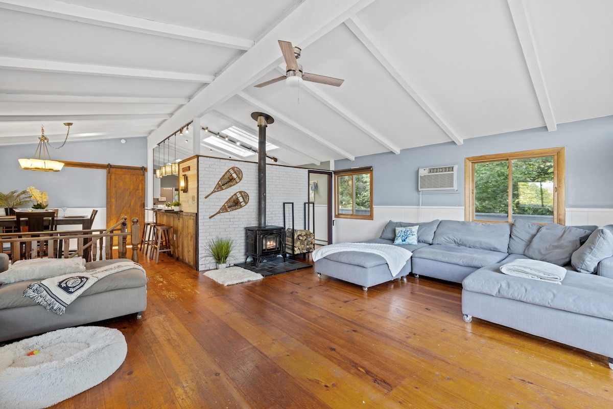 An open concept living area showcases high, beamed ceilings and large windows allowing natural light to illuminate the space. A cozy sectional couch is positioned near a wood-burning stove, with a dining area visible in the background, featuring a large table and chandelier.