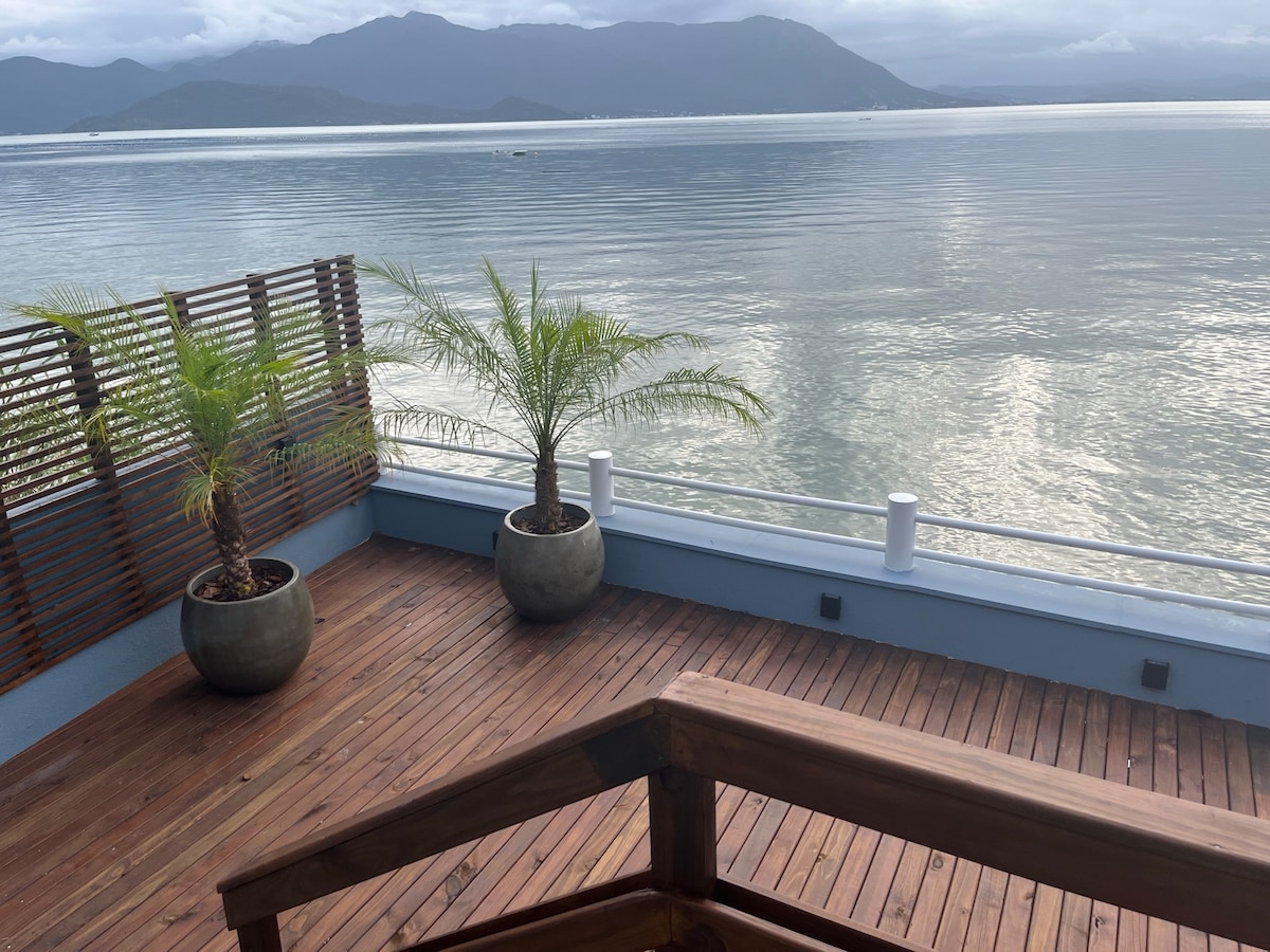The outdoor deck features a wooden surface, complemented by two potted palm trees. A railing provides a clear view of the tranquil water and distant mountains, with soft waves reflecting the sky above. The serene scene invites relaxation and appreciation of the natural surroundings.