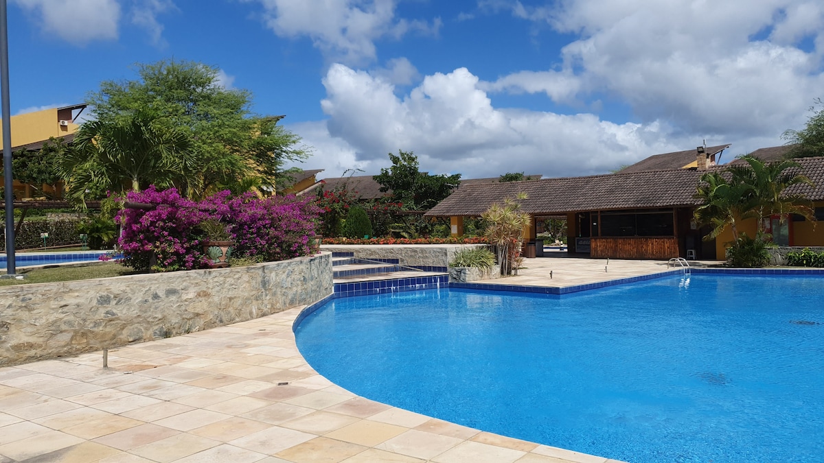 A serene outdoor pool area is surrounded by lush greenery and vibrant flowering plants. The pool features clear blue water and a stone border, with a spacious patio area visible. Buildings with traditional roofs can be seen in the background under a partly cloudy sky.