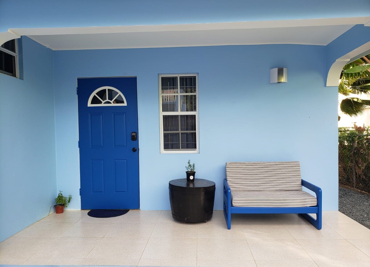 A welcoming entrance features a bright blue door, complemented by a light blue exterior. A sleek outdoor bench and a small round table are positioned on the tiled porch, creating an inviting spot. A small plant is placed on the table, adding a touch of greenery.