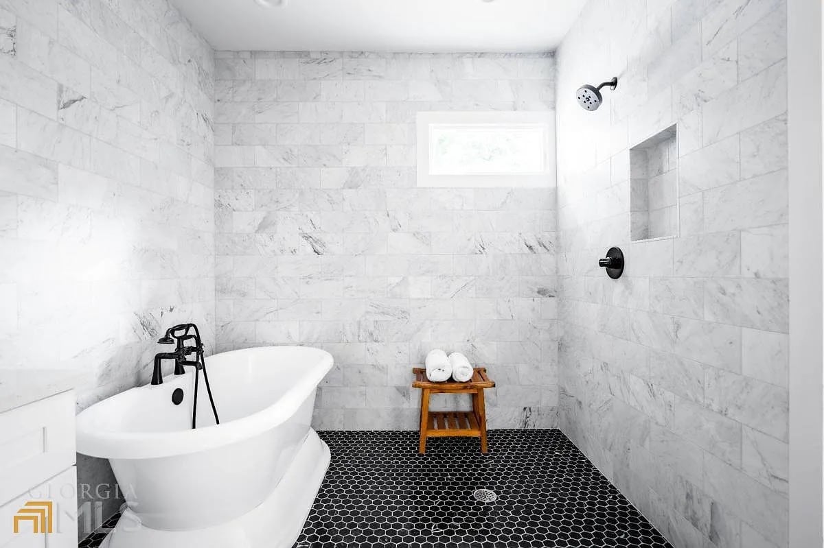 A modern bathroom features a standalone tub alongside a spacious walk-in shower. The walls are adorned with white marble tiles, and a large window allows natural light to enter. A wooden stand holds neatly rolled towels, complemented by black hexagonal floor tiles.