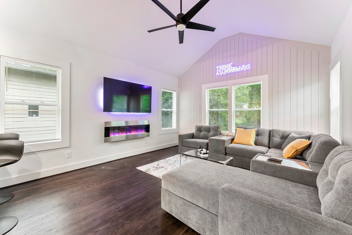 A spacious living area features a comfortable sectional sofa and a sleek coffee table, with a wall-mounted television providing entertainment. Large windows invite natural light, while a contemporary ceiling fan adds to the room's airy ambiance. A soft area rug lies underfoot.