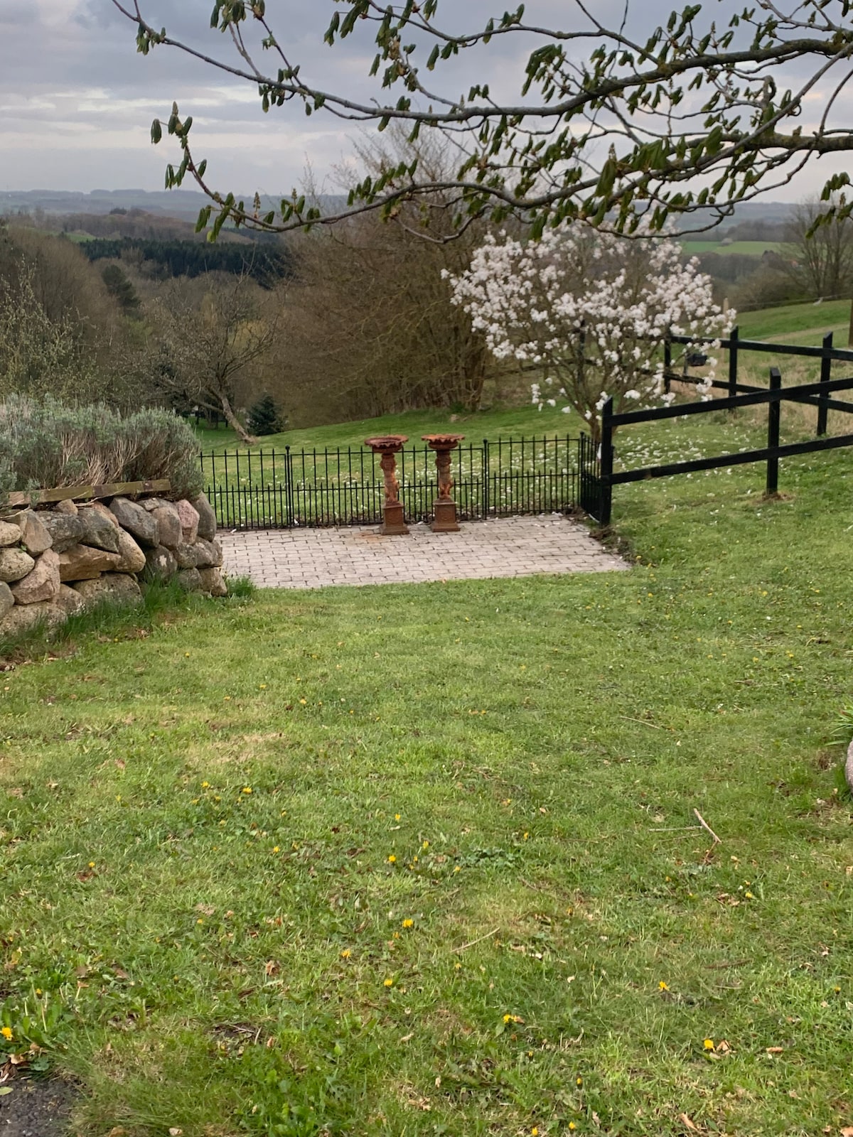 A serene outdoor area features a neatly maintained lawn bordered by a stone wall. A pair of decorative columns stand at the entrance of a fenced pathway, which leads to a scenic view of greenery and tree line in the background.