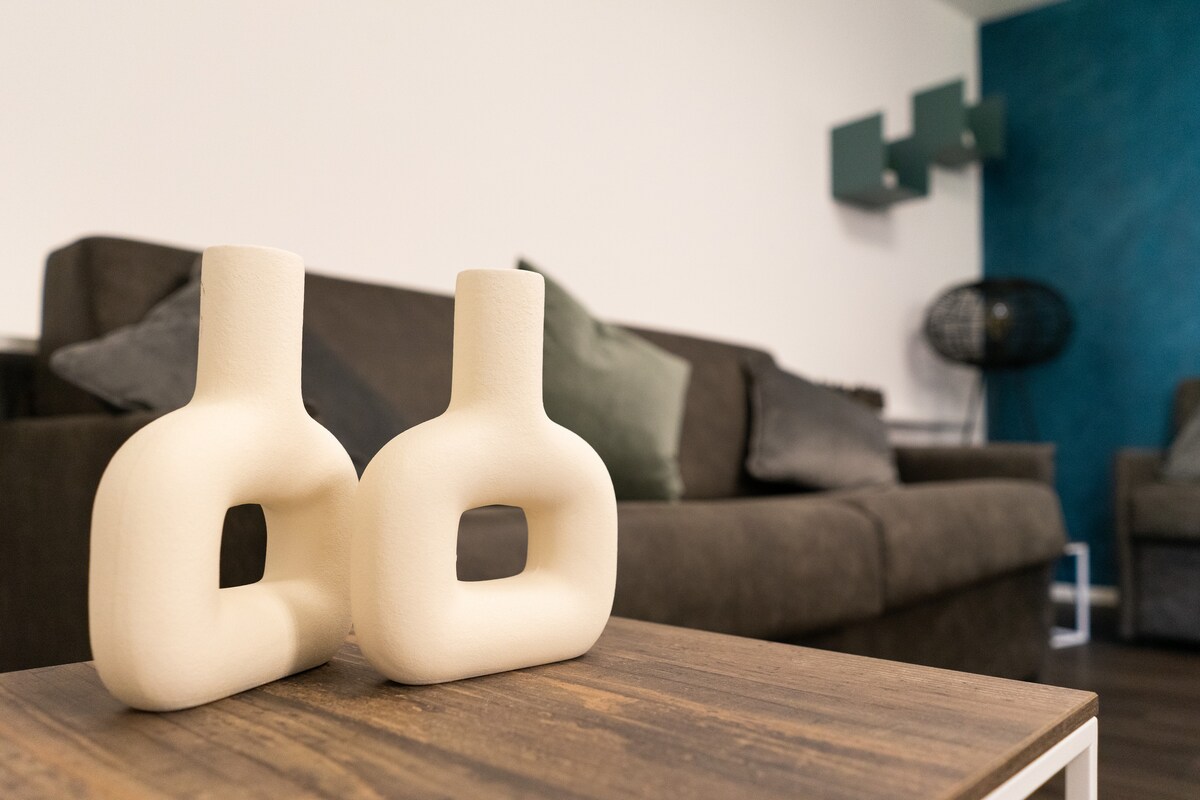 Two sculptural vases are placed on a wooden coffee table. A soft, dark sofa is visible in the background, adorned with cushions in varied hues, while a textured blue wall adds character to the space. A stylish chair can be seen to the side.