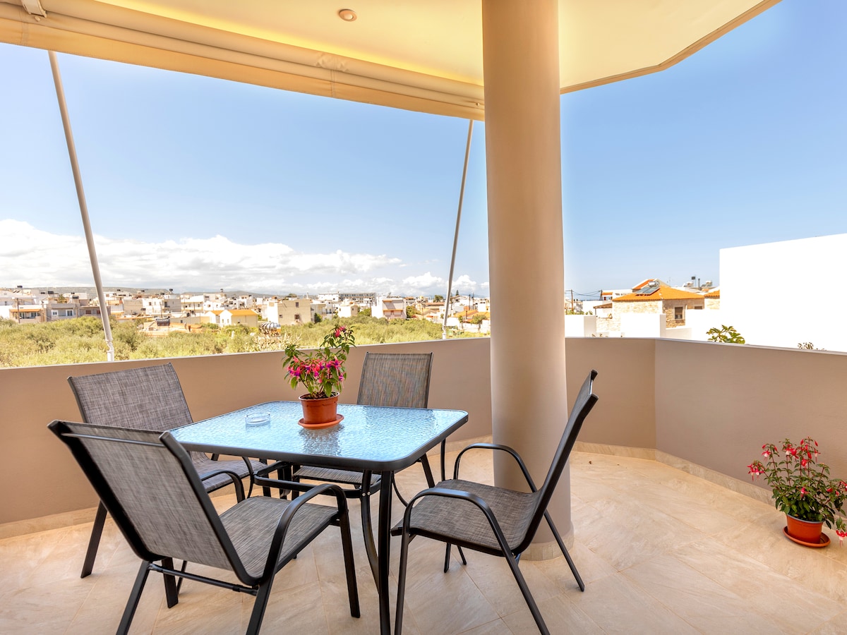 A spacious balcony is showcased, furnished with a glass-top table surrounded by four chairs. Potted plants add a touch of greenery, while expansive views of the surrounding area and distant hills can be seen under a clear blue sky.