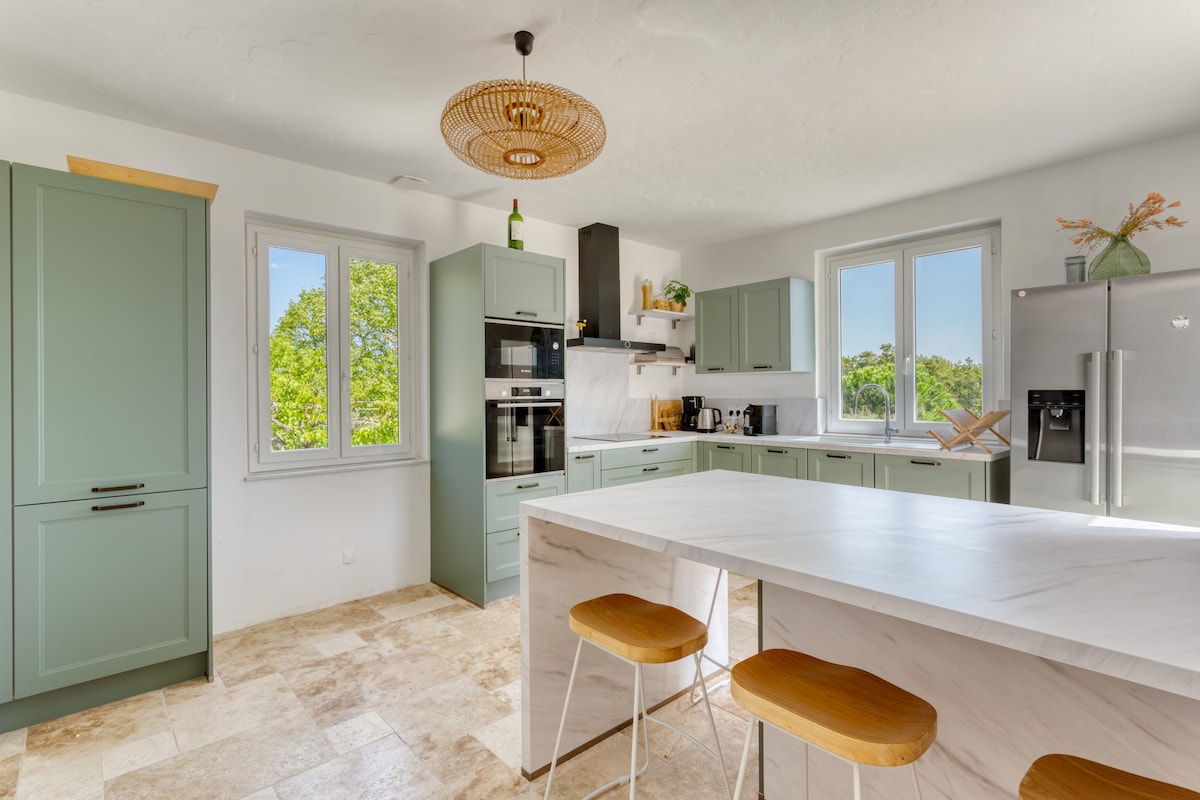 A modern kitchen features green cabinetry and a large marble island. Natural light enters through two windows, highlighting the spacious layout. Equipped with built-in appliances, the kitchen offers a minimalist design with bar stools positioned around the island.