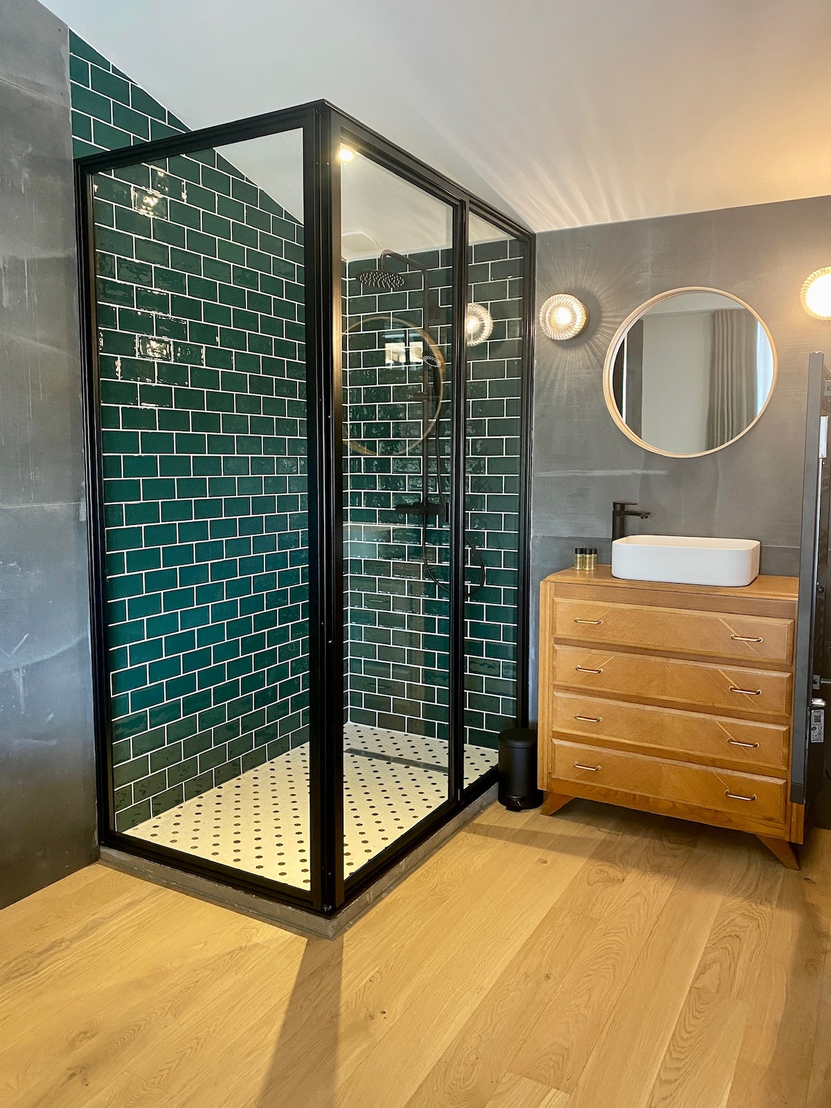 A modern bathroom features a glass shower enclosure with dark metal framing, displaying green subway tiles. A circular mirror hangs above a white sink on a wooden vanity, complemented by soft lighting fixtures. The floor is covered in light wooden planks, enhancing the sleek design.