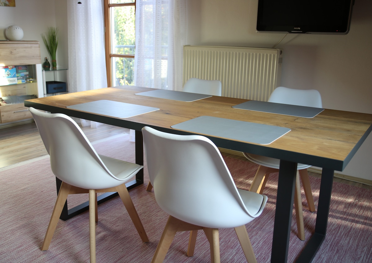 A modern dining area features a wooden table with natural grain, surrounded by four white chairs with wooden legs. Light streams through large windows, illuminating minimalist placemats on the table. A radiator and a television are visible in the background.