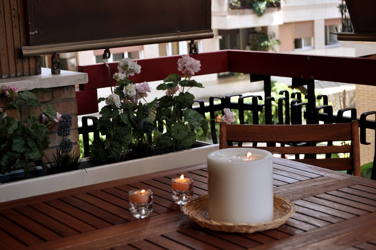 A wooden table is set on a balcony, featuring a large white candle and smaller glass candles. Green plants with pink flowers are visible in the background, adding a touch of nature to the outdoor space.