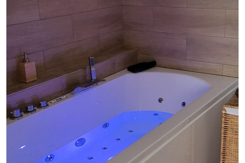 A modern jacuzzi tub is set against a backdrop of wooden tiles, featuring soft blue lighting that enhances the relaxing atmosphere. Controls and fixtures are neatly arranged, and a towel is placed on the edge, ensuring convenience for guests.